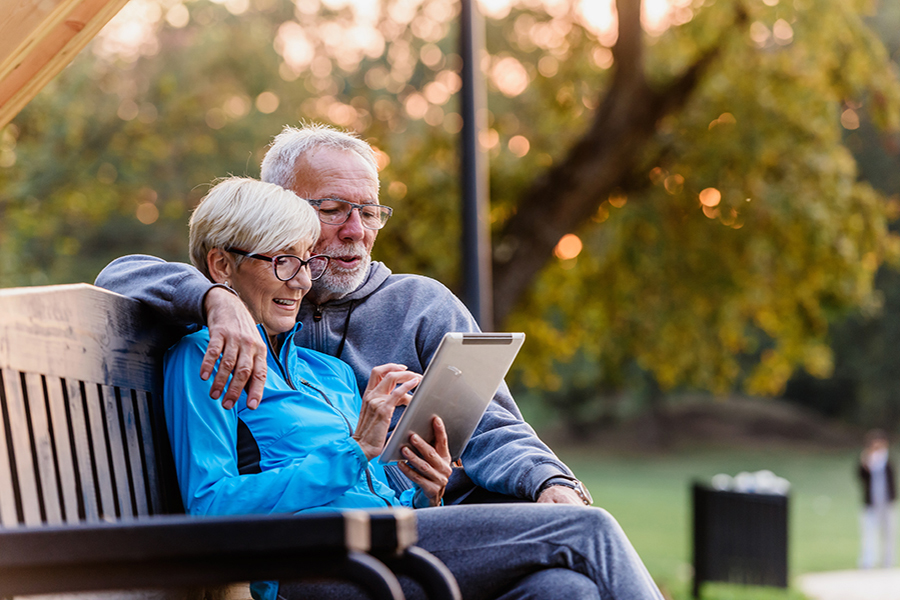 Smiling senior active couple sitting on the bench looking at tab
